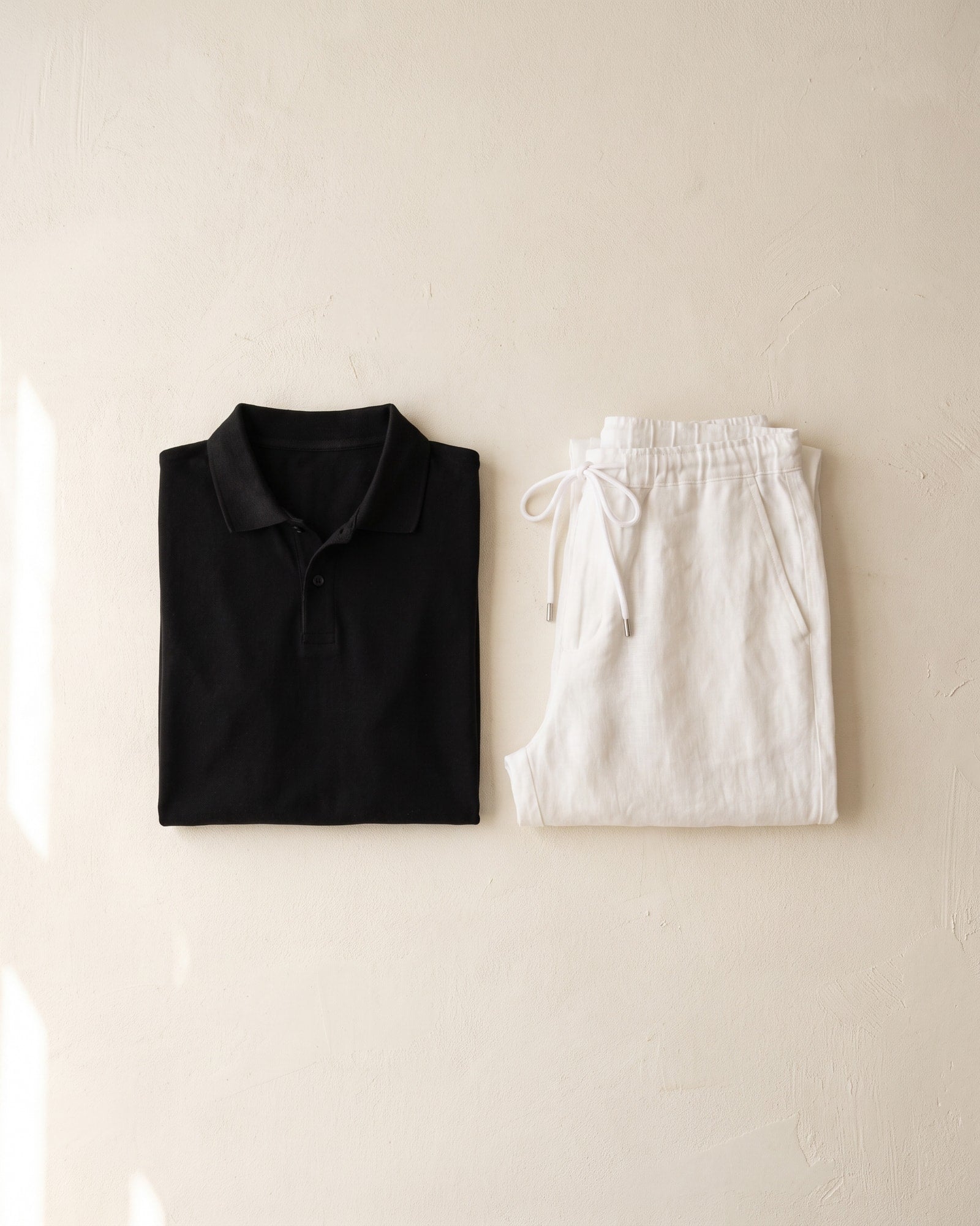 Folded Cole Jackson black polo and white linen trousers on a warm plaster wall